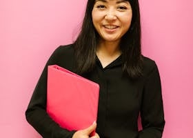 Asian woman in black attire smiles holding a pink binder against a pink background.